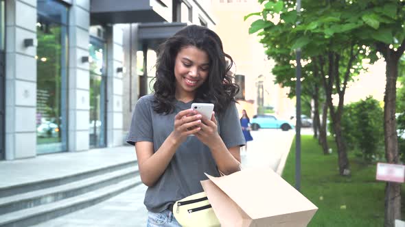 Happy Young African Woman Standing on Street Holding Shopping Bags and Phone