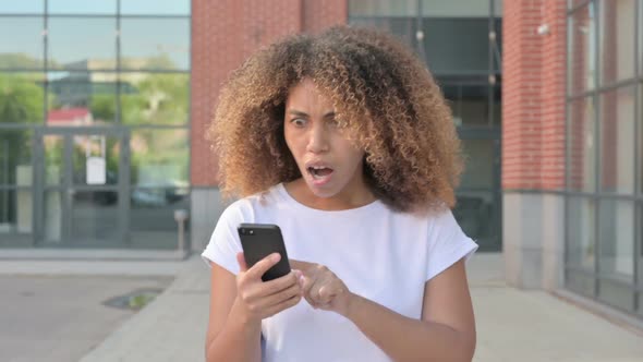 African Woman Reacting to Loss on Smartphone While Walking Outside