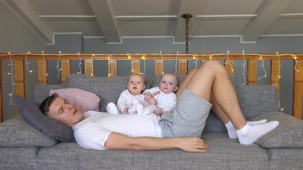 Tired Father Sleeping On Sofa With His Twin Babies At Home. Baby And Father Concept.