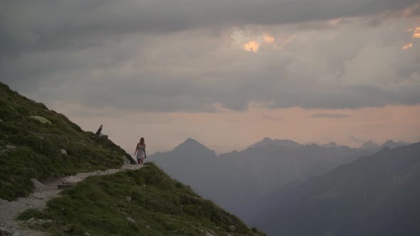 White girl in a white dress appears behind a mountain and walks on an exposed mountain path towards