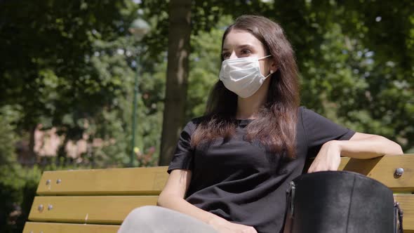 A Young Turkish Woman in a Face Mask Looks Around As She Sits on a Bench in a Park