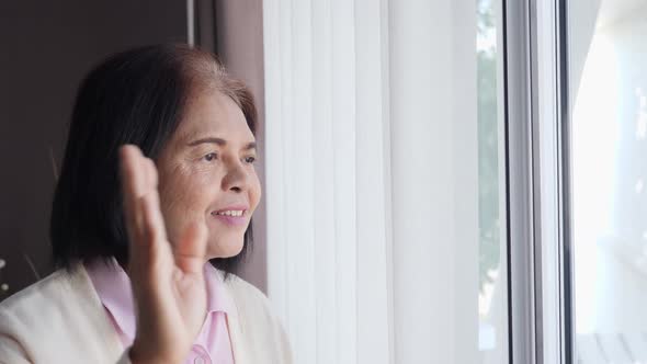 Happy senior Asian woman looking outside windows, waving hand, and smiling