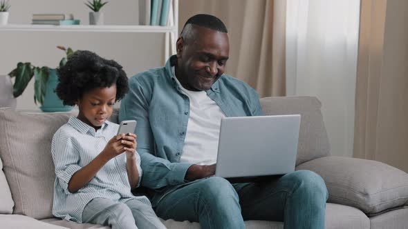 Happy African American Family Relaxing on Sofa at Home Man Typing on Laptop Kid Girl Playing on