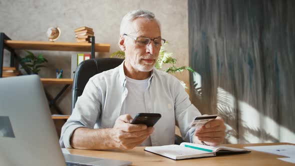 Senior Man Holding Credit Card and Entering Its Number Into Mobile Phone Shopping Online Working at