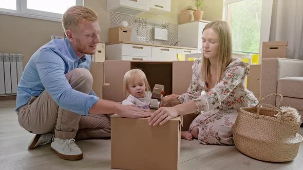 Parents and Two Little Daughters Sit on Floor and Unpack Family Suitcase in New House