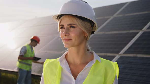 Beautiful Female Solar Engineer Standing Near the Panels and a Young Specialist in the Background