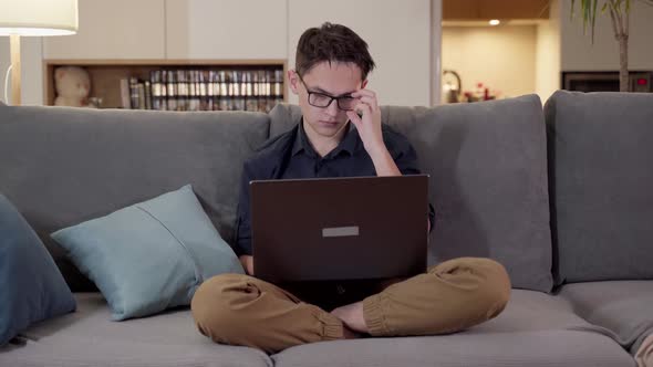 A Young Guy with Glasses Sits on a Sofa with a Laptop