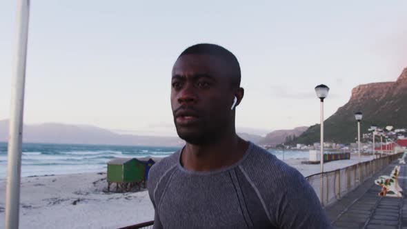 Focused african american man exercising outdoors, running by seaside with wireless earphones on