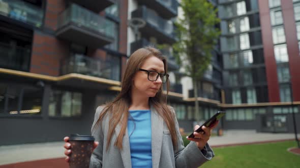 Formally Dressed Woman Walking Down the Street in a Business District with Coffee in Hand and Using