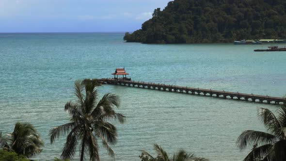 A pier with a small red roof at the end, in the ocean with palm trees Slowmo