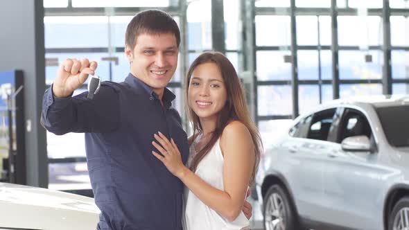 Happy Caucasian Couple Hugging in Front of New Car, Holding the Keys, at the Car Dealership.