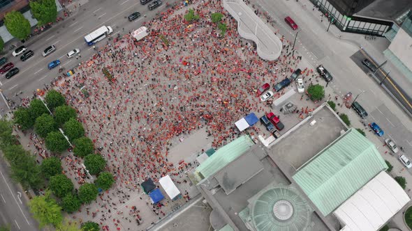 Aerial view of an event in a downtown public square, wide drone overhead. Busy city traffic passes a