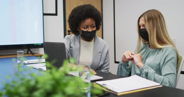 Two diverse businesswomen wearing face masks working together using laptop at desk in office