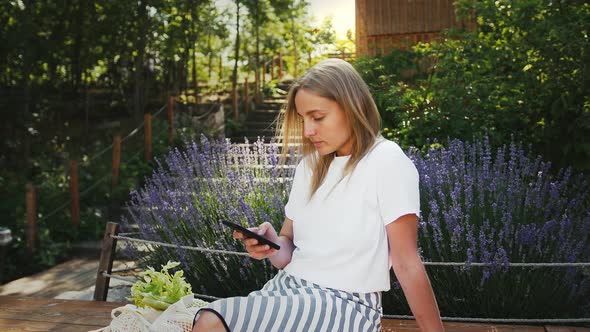 Young Woman is Using Her Smartphone While Sitting on a Bench of City Park