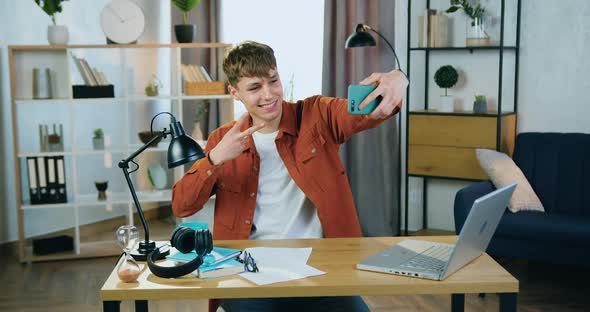 Guy Making Selfie on Smartphone with Fingers v-sign Sitting at the Table in Cozy Room at Home