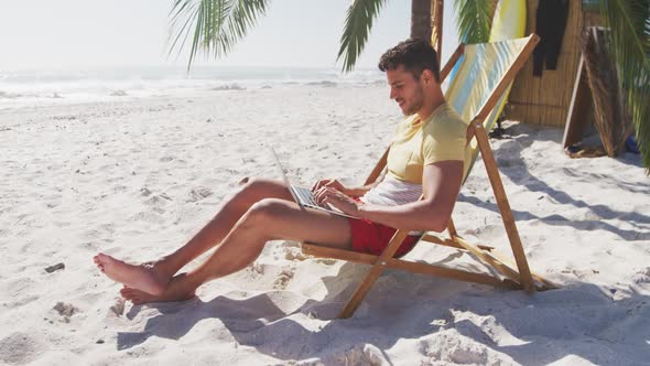 Caucasian man sitting on a sunbed and using his laptop on the beach