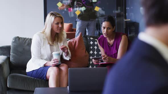 Diverse businesswomen sitting using laptop drinking coffee in office lounge