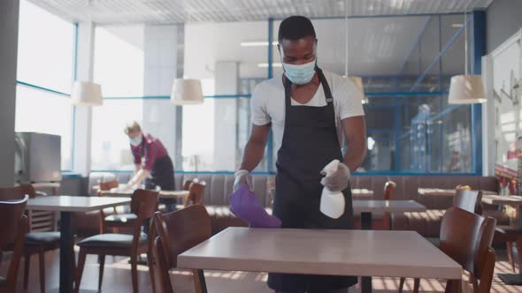 AfricanAmerican Waiter in Safety Mask and Gloves Clean Table in Cafe