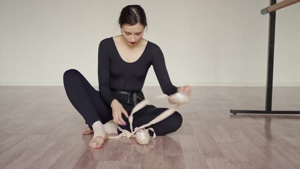 a Beautiful Dancer in Black Clothes Sits Next to a Ballet Barre on the Floor and Puts on Pointe