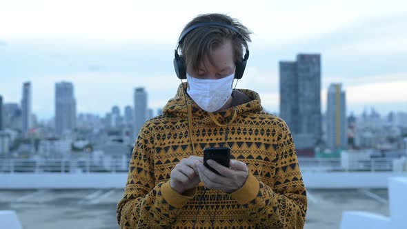 Young Man in Hoodie Using Phone While Listening To Music with Mask Against View of the City