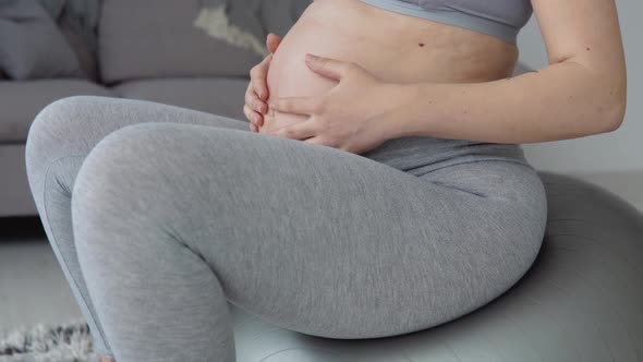 A Pregnant Woman in Sportswear Sits on a Fitball and Holds Her Hands on Her Stomach