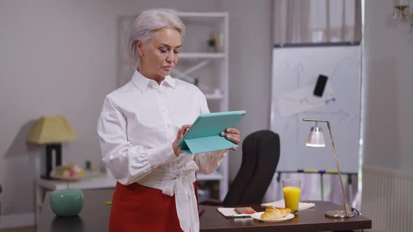 Concentrated Slim Gorgeous Mature Businesswoman Standing in Office Analyzing Emarket on Tablet