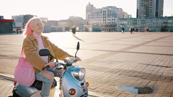 Young Woman with Pink Hair Enjoying Ride on Scooter Riding Along City Square in Sunny Day Tracking