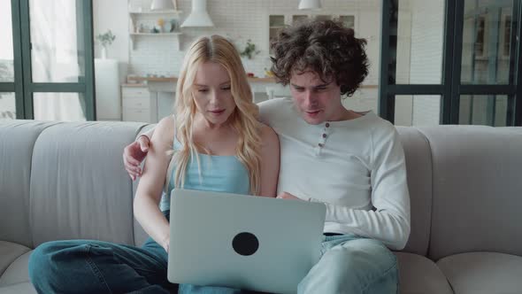 Young Woman Sitting in Couch with Computer on Laps with Smiling Pretty Boyfriend Sitting Beside