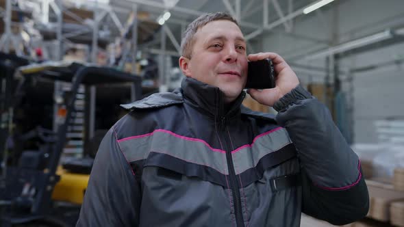 Cheerful Adult Caucasian Man in Uniform Talking on Phone Standing in Warehouse Indoors