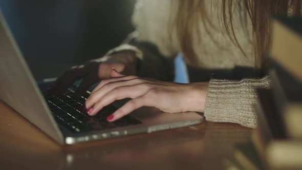 View of Female Hands Typing on Laptop Keyboard at Desk with Books on Dim Light