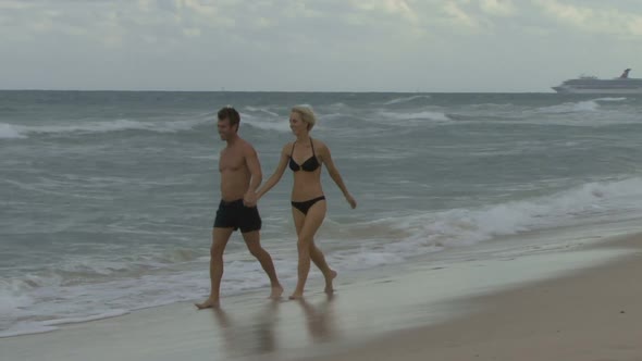 Couple holding hands walking along a beach with a cruise ship in the background