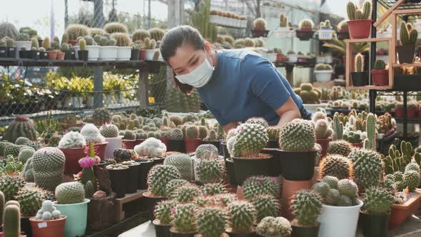 A masked Asian woman examines the cacti on a table in a plant nursery and picks up one pot. Slow mot