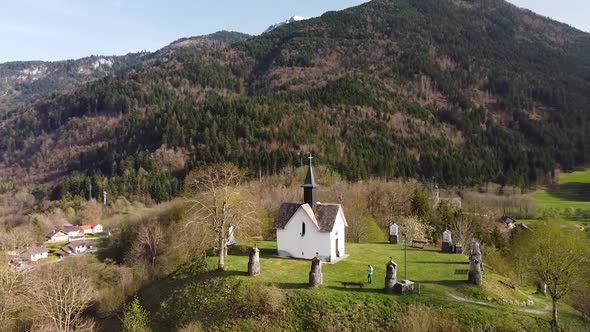 Drone Shot (facing forwards, rotating) of a Chapel on top of a Hill in front of Alpine Mountains