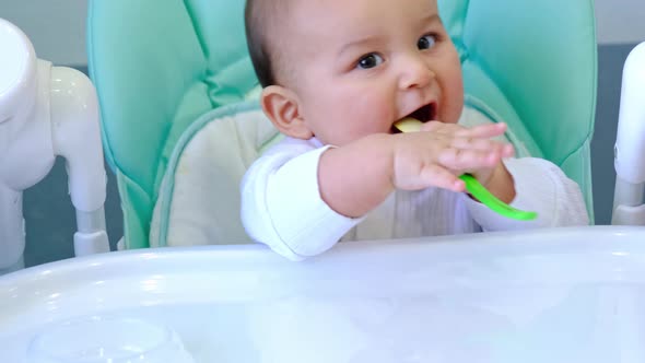 A hungry baby is gnawing on a plastic spoon at the table on a high chair. Teething, whims, itchy gum