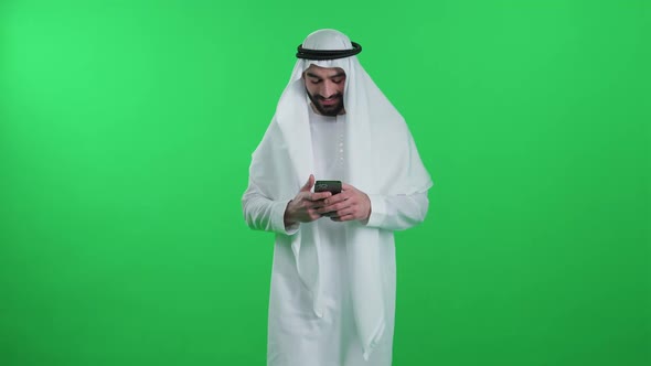 An Arab Man in a White Robe on the Green Background Wearing Kandura Walks Using a Smartphone and