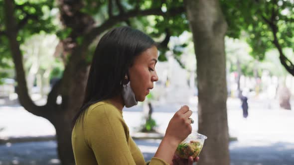 African american woman wearing face mask eating salad in sity park