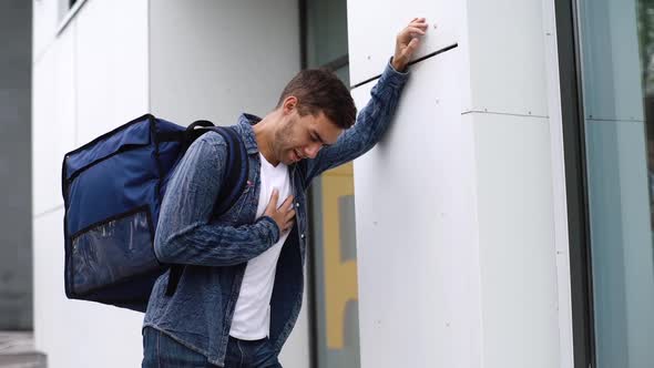 Exhausted Food Delivery Man with Large Thermo Backpack Stopping By Wall and Holds Heart Breathing