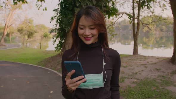 Portrait of Happy Thai Young Woman Wearing Protective Face Medical Mask Standing in Park at Sunset