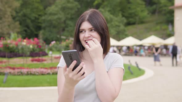 A Young Caucasian Woman Works on a Smartphone with a Smile in a City Park on a Sunny Day