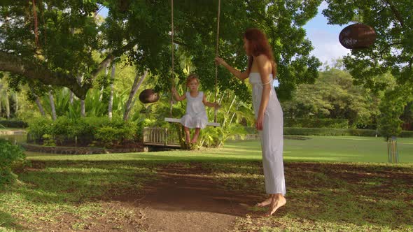 Mom and Daughter in White Dresses Spend the Weekend in a Green Park with a Swing