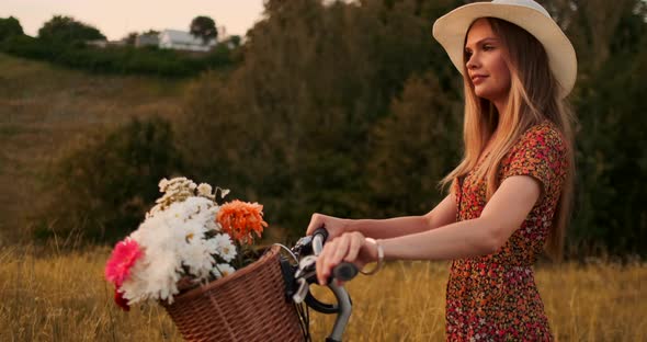 Young Smiling Blonde in Hat and Dress Walking in Dress with Bike and Flowers in Basket