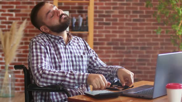 Tired Hipster Businessman is Sitting on Wheelchair at Table with Laptop and Rubbing His Eyes