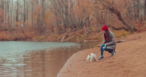 Girl Sits Near the Shore with a Small Dog, Treats the Puppy with a Treat. Walk By the River in the