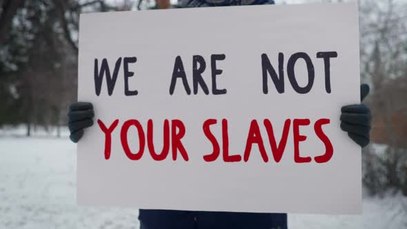 A Protester is Standing in a Park with a Sign We Are Not Your Slaves. Picket.