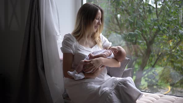 Happy Smiling Caucasian Woman Sitting on Windowsill Shaking Newborn Baby in Slow Motion Looking Out
