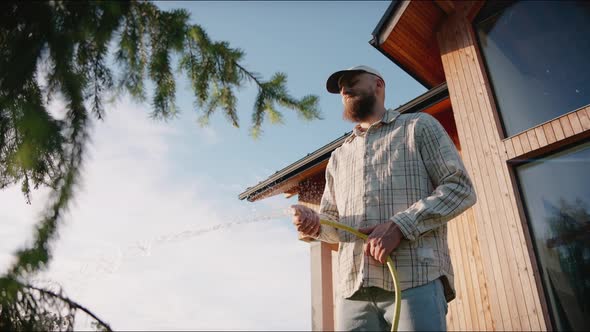 Man Waters Plants and Trees with a Hose at Summer Sunset in the Backyard