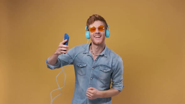 Young Man Listening Music on Mobile Phone Application with Headphones and Dancing Isolated on Yellow