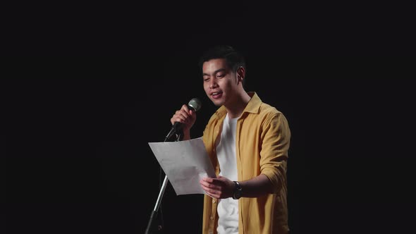 Side View Of Asian Man Singer Holding White Paper And Singing Into Microphone On Black Background