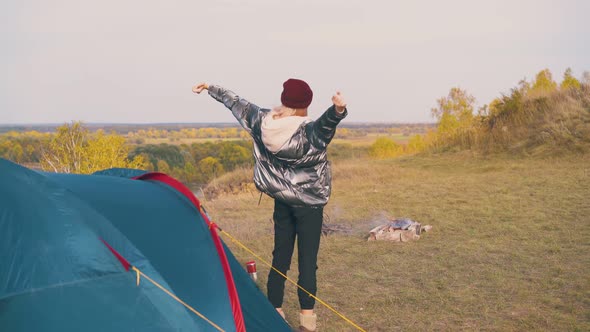 Girl Tourist Stretches at Tent and Bonfire on River Bank