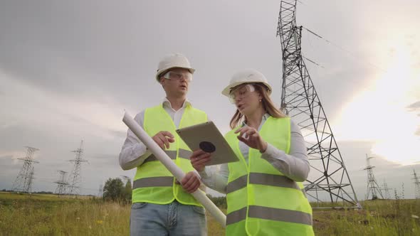 Two Electricians Work Together Standing in the Field Near Electricity Transmission Line in Helmets